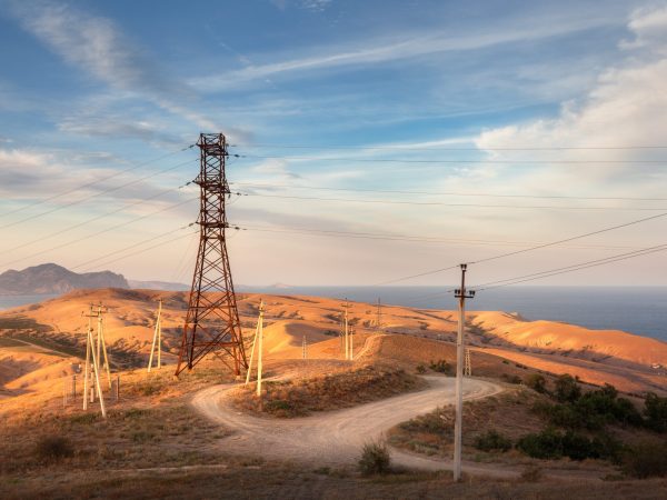 High voltage tower, road, hills with yellow grass in mountains on the background of colorful sky with clouds at sunset.  Electricity pylon system in the hill. Summer evening. Industrial landscape